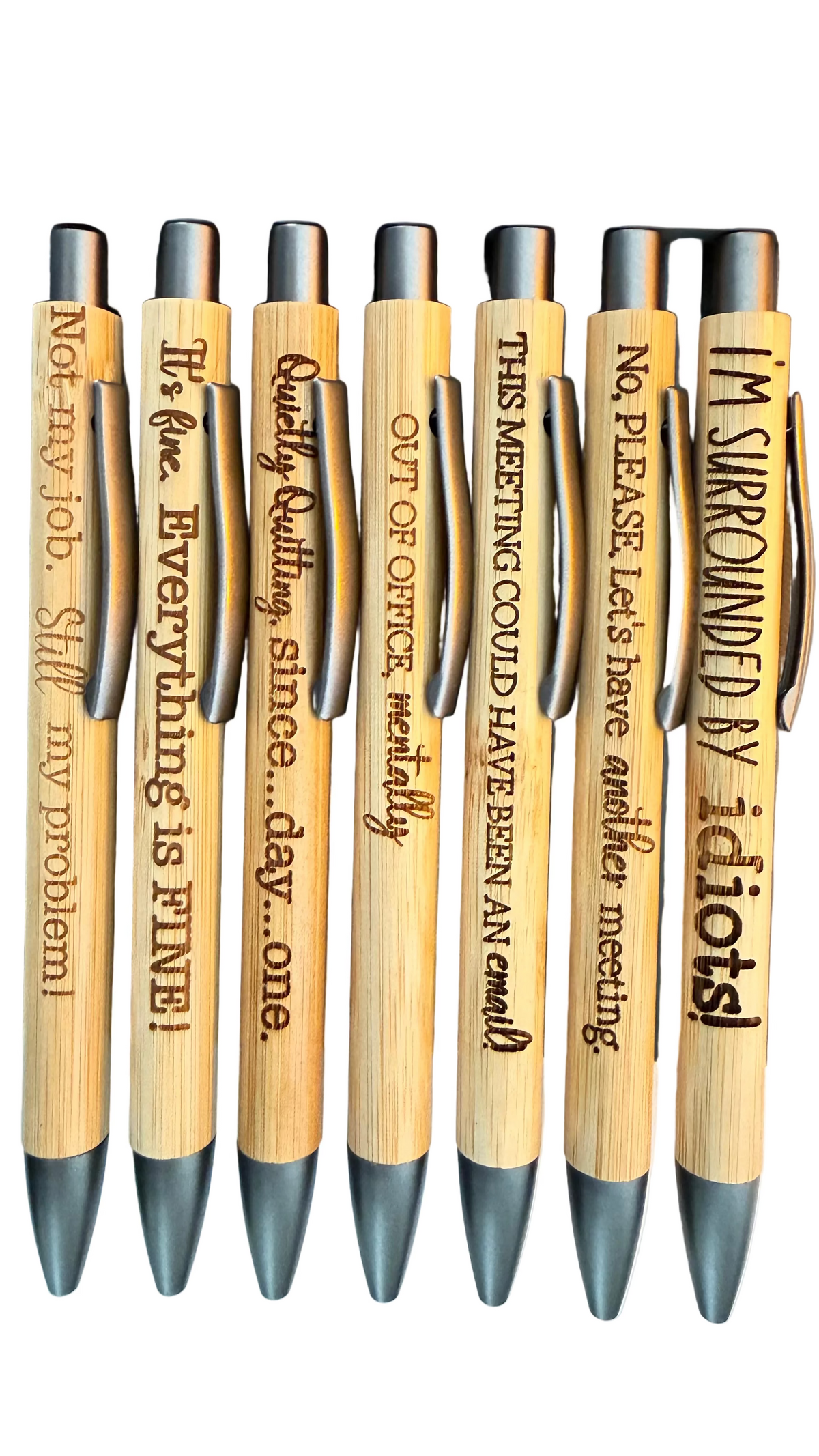 A set of eight bamboo pens with black metal tips, arranged in a row inside a black tray. Each pen is engraved with a humorous quote in black lettering.