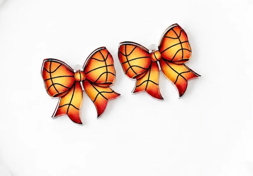 A pair of bow-shaped earrings designed to look like basketballs, featuring a vibrant orange and yellow gradient with black curved lines mimicking a basketball's seam pattern, set against a plain white background.