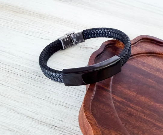 A black braided leather bracelet with a glossy black metal ID plate and silver-toned clasp, resting on a dark wooden tray over a light wood surface.