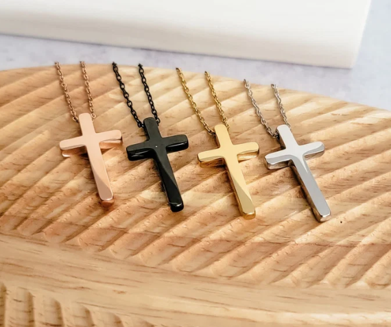 Four cross-shaped pendant necklaces in rose gold, black, yellow gold, and silver, displayed on a carved wooden surface.