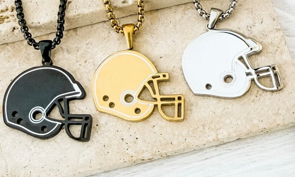 Three football helmet pendant necklaces displayed on a light stone surface, shown in black, gold, and silver finishes.