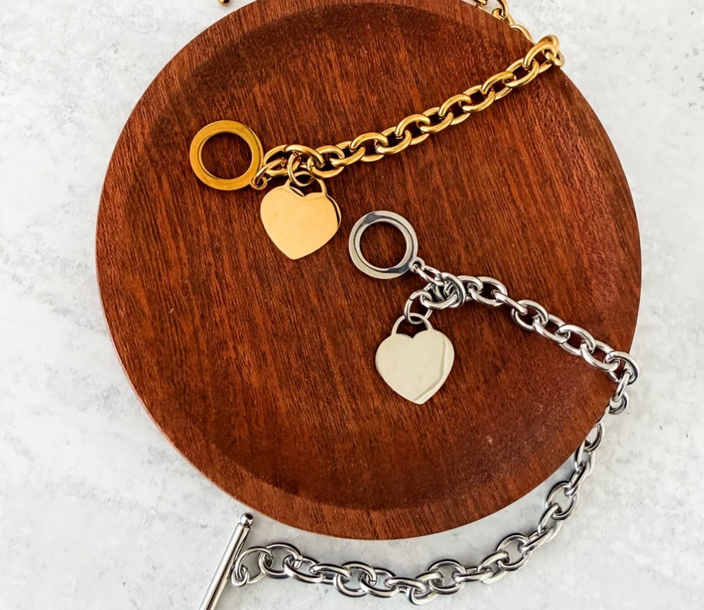 A gold and silver chain bracelet with heart charms, displayed on a round wooden stand over a light marble surface.