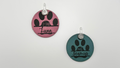 Two round retractable dog leash collars on a light gray background, one in pink with black paw prints and the name 'Luna', and one in teal with black paw prints and the name 'Beverly'.