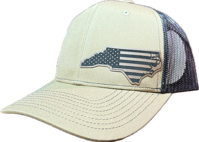 A beige trucker cap with a dark mesh back and a gray embroidered patch of the state of North Carolina filled with a distressed American flag design.