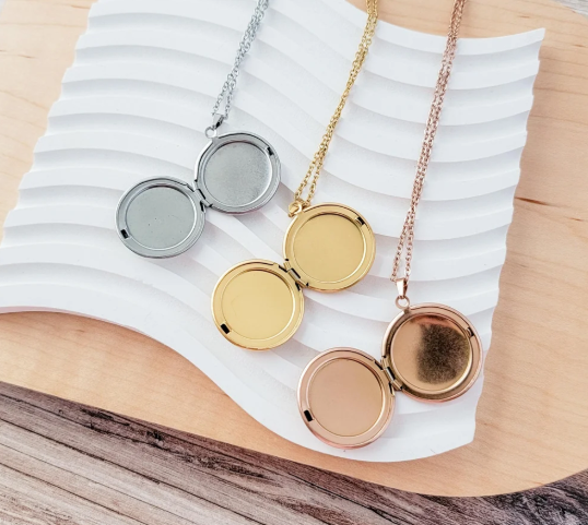 Three open locket necklaces in silver, gold, and rose gold are displayed on a white wavy dish over a light wood surface.