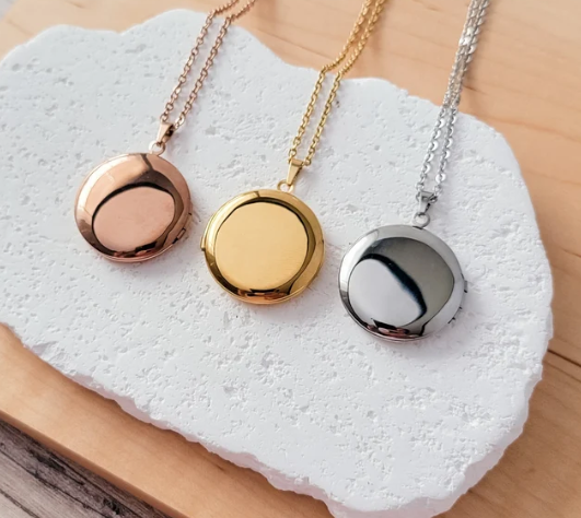 Three round locket necklaces in rose gold, yellow gold, and silver, displayed on a white textured stone tray over a light wood surface.