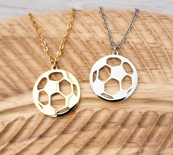 Two soccer ball cutout pendant necklaces, one gold and one silver, displayed on a wooden surface.