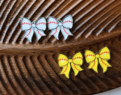 Two pairs of bow-shaped earrings designed like baseballs, one pair in silver and white with red stitching, and one pair in yellow and white with red stitching, displayed on a dark wood background.