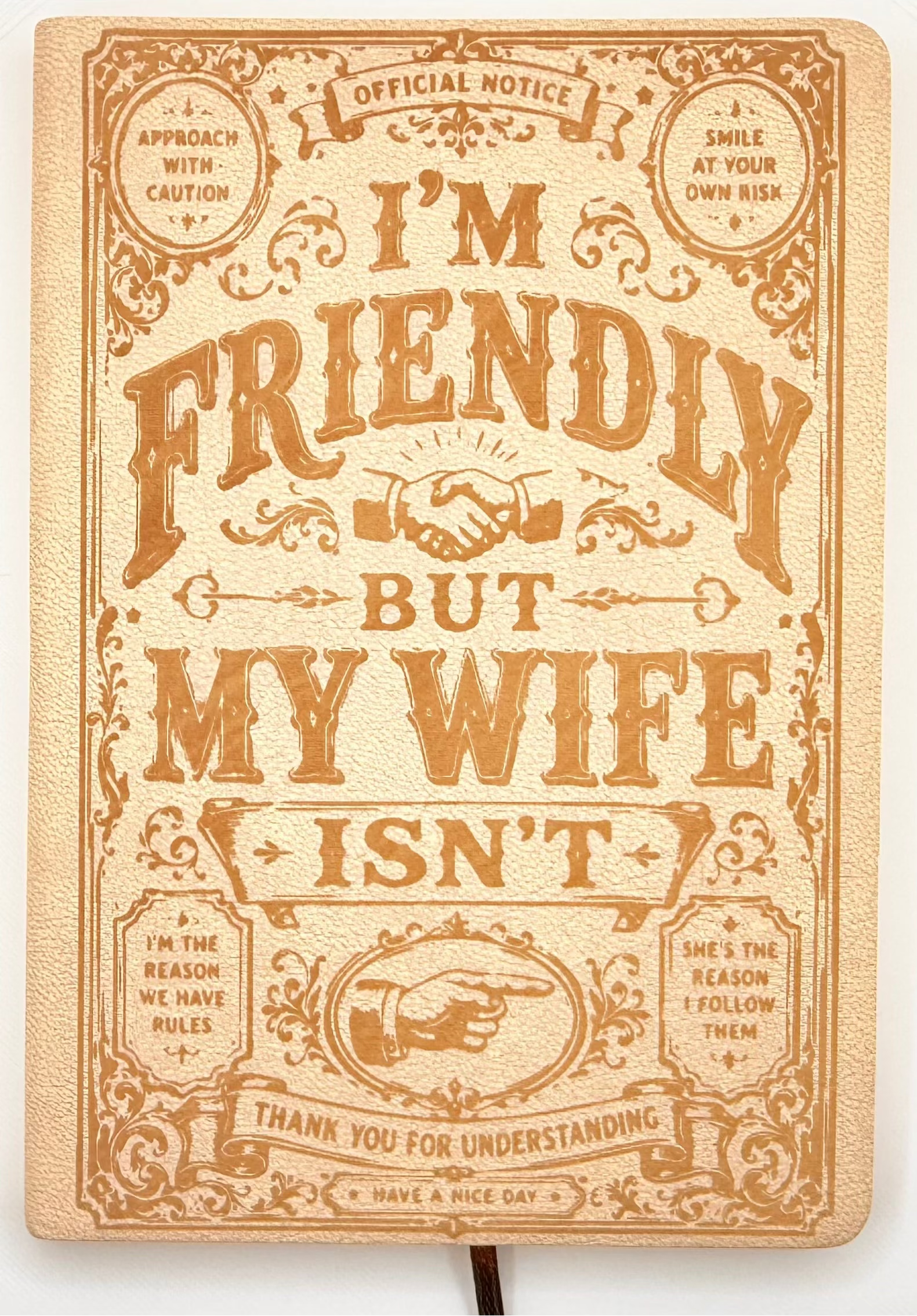 A square decorative sign or plaque with ornate vintage-style typography reading 'I'M FRIENDLY BUT MY WIFE ISN'T' in warm tan lettering on a cream background, photographed against a plain white wall.