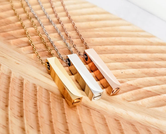 Three minimalist bar pendant necklaces in gold, silver, and rose gold finishes, displayed on a textured light wood background.