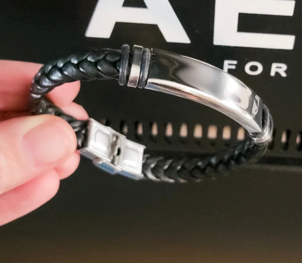 A close-up of a hand holding a black braided leather bracelet with a polished silver-tone metal ID plate and clasp, set against a dark background with white lettering.