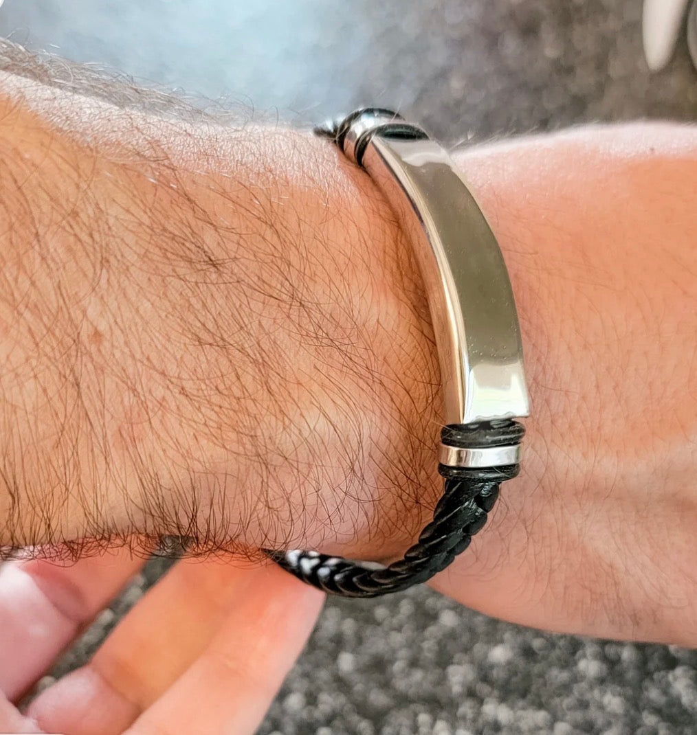 A close-up of a man's wrist wearing a black braided leather bracelet with a polished silver-tone ID plate.