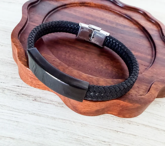 A black braided leather bracelet with a polished black bar and silver-toned clasp, displayed on a scalloped wooden tray over a light wood surface.