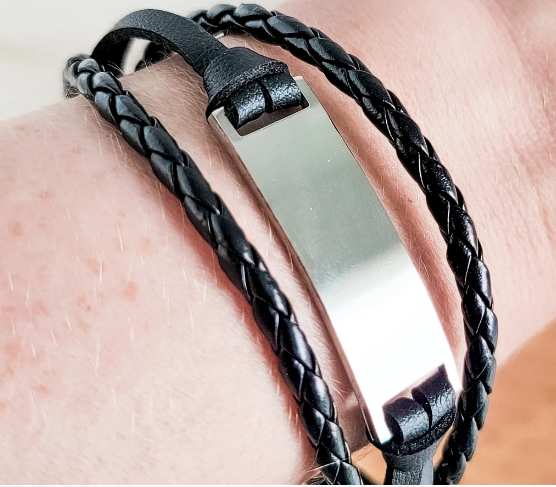 A close-up of a black leather wrap bracelet with a polished silver-toned metal ID plate, worn on a person's wrist.