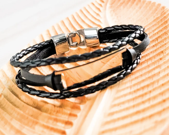 A black braided and woven leather bracelet with a polished silver-toned metal clasp, resting on a light wood grain surface.